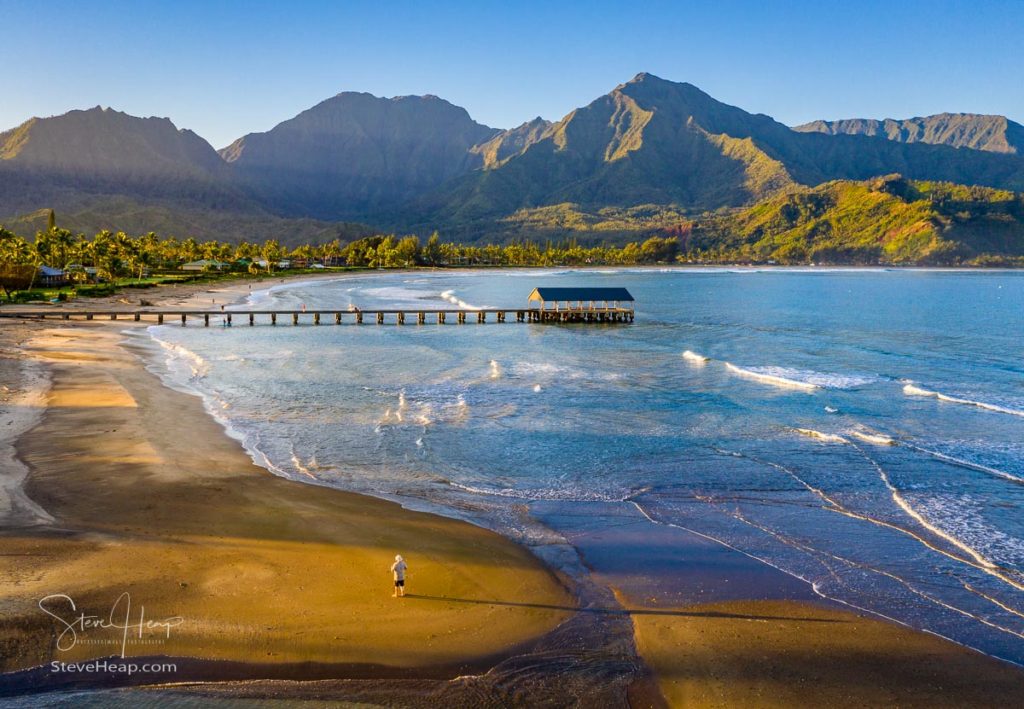 Aerial image at sunrise off the coast over Hanalei Bay and pier on Hawaiian island of Kauai with a man standing alone on the beach. Prints here