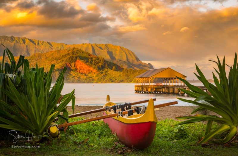 Red and yellow hawaiian canoe with outrigger on the beach at Hanalei pier at dawn as the sun lights the sky over Na Pali mountains