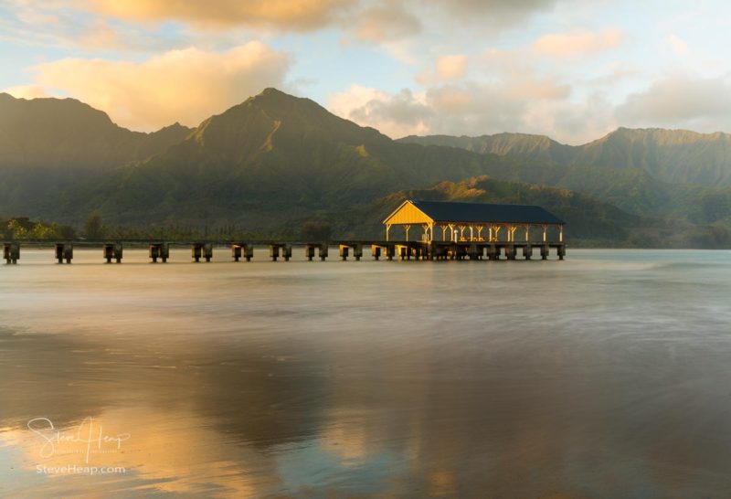 Rising sun illuminates the peaks of Na Pali mountains over the calm bay and Hanalei Pier in long exposure photo