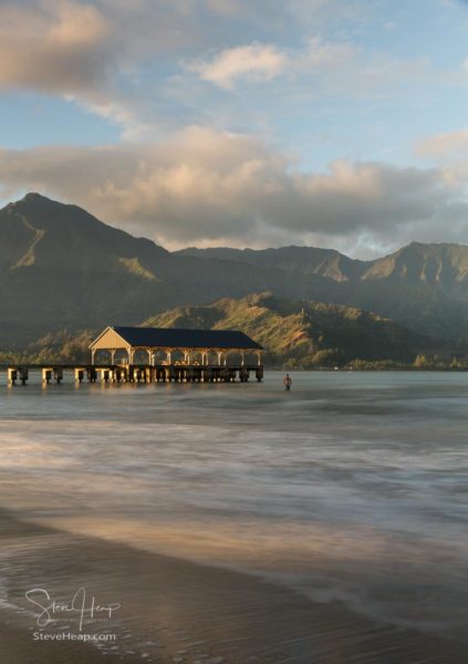 Swimmer stands in the ocean as the rising sun illuminates the peaks of Na Pali mountains over the calm bay and Hanalei Pier