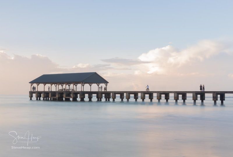 Tourists and sightseers watch sunrise from Hanalei Pier in Kauai Hawaii with long exposure blurred motion ocean waves