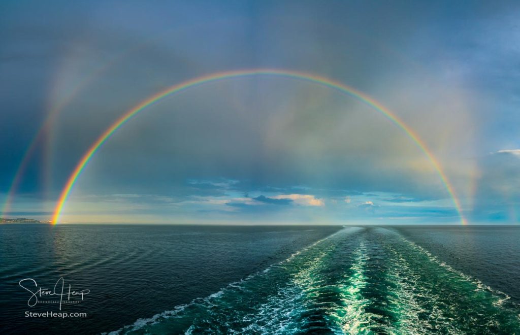 Colorful and dramatic double rainbow forms a full arch over the wake of a departing cruise ship at sea. Prints available in my store