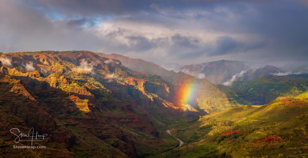 A dramatic rainbow peeps through the storm clouds over Waimea Canyon in this panorama of the Hawaiian island of Kauai. Prints in my store