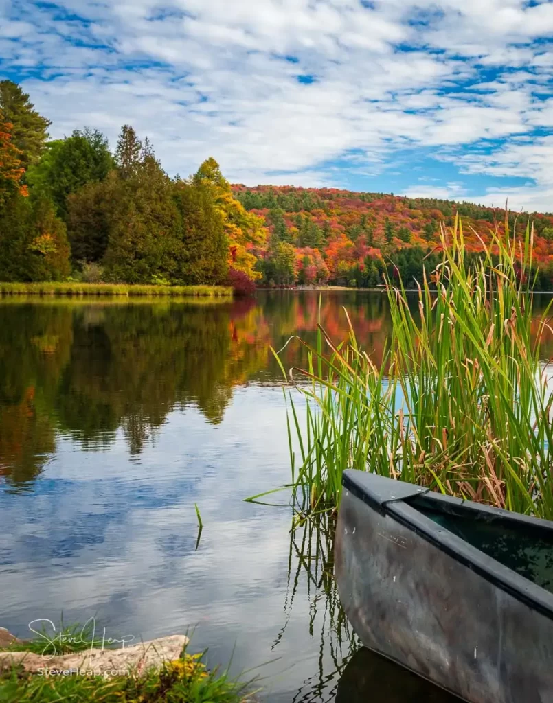 An old canoe leaving the reeds to head out into a calm lake reflecting the fall colors of the trees in Vermont. Prints in my online store
