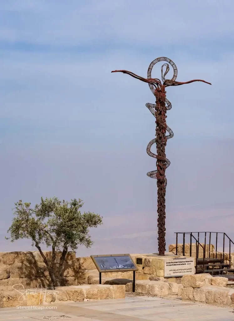 A sculpture of the Brazen Serpent cross, created by Paolo Fantoni, situated on a hilltop in Jordan, with an olive tree and informational plaque nearby.