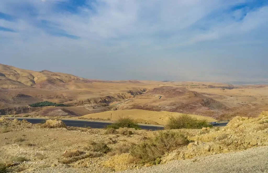 A panoramic view of a barren landscape in Jordan, showcasing rolling hills and winding roads against a backdrop of cloudy blue skies.