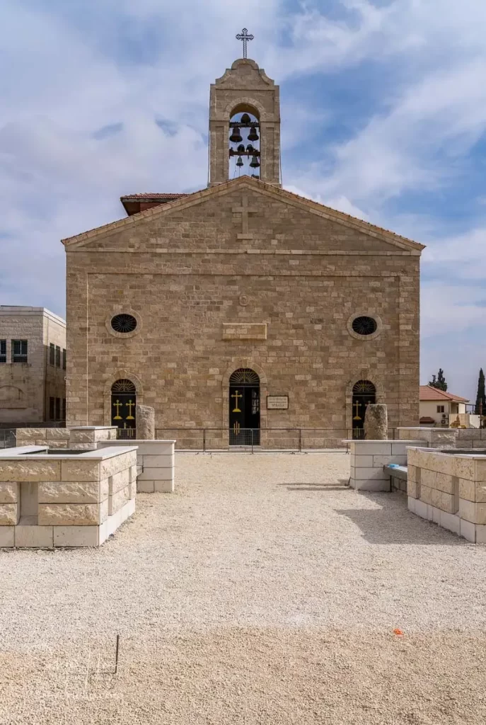Exterior view of the Moses Memorial Church at Mount Nebo, featuring a stone facade with a bell tower and cross on top, surrounded by a gravel plaza.