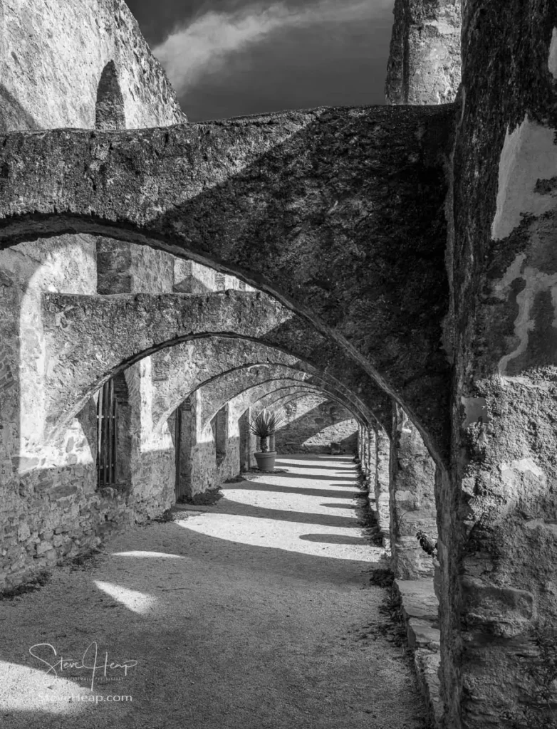 Black and white photograph of a long stone colonnade at the San Jose mission, featuring repeating arches and dramatic afternoon shadows.