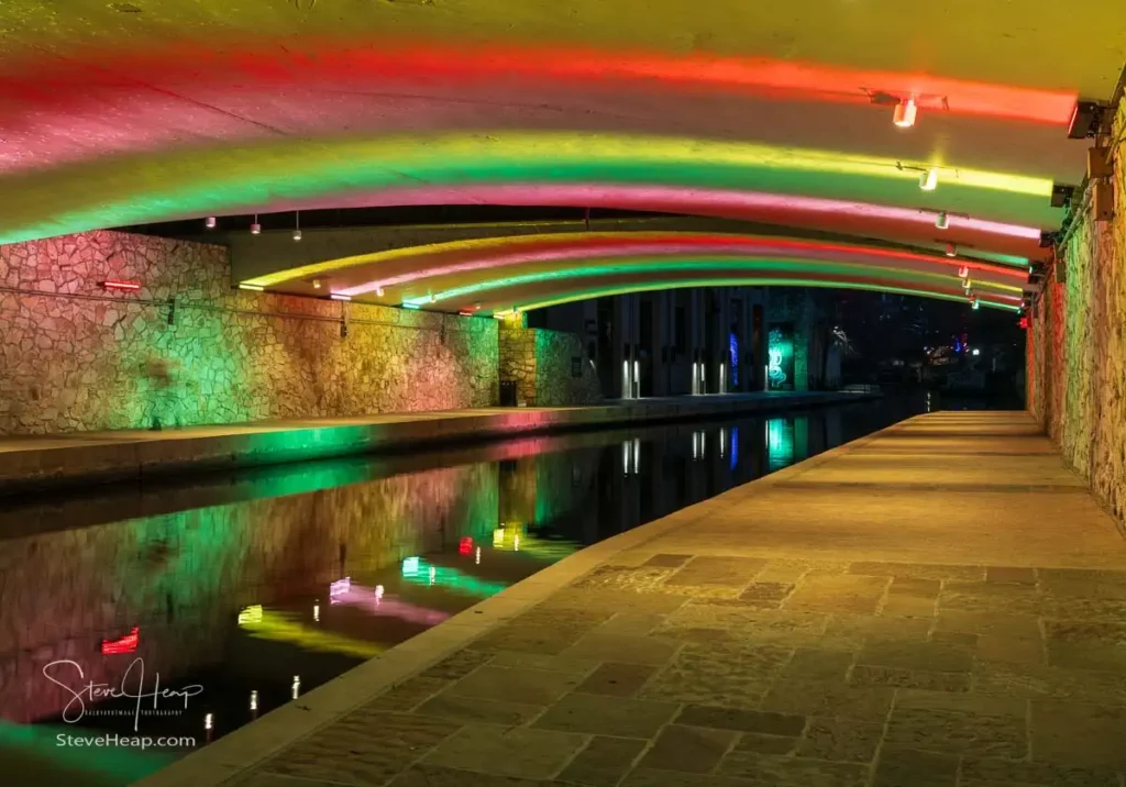 Vibrant green and red LED lights illuminating the brickwork and water underneath a bridge on the San Antonio River Walk at dawn.