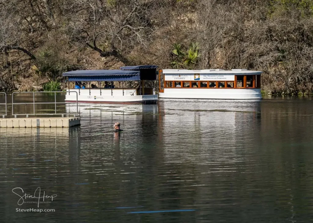 Wide landscape shot of a glass-bottom boat resting on clear blue water, reflecting the surrounding trees at the San Marcos springs.
