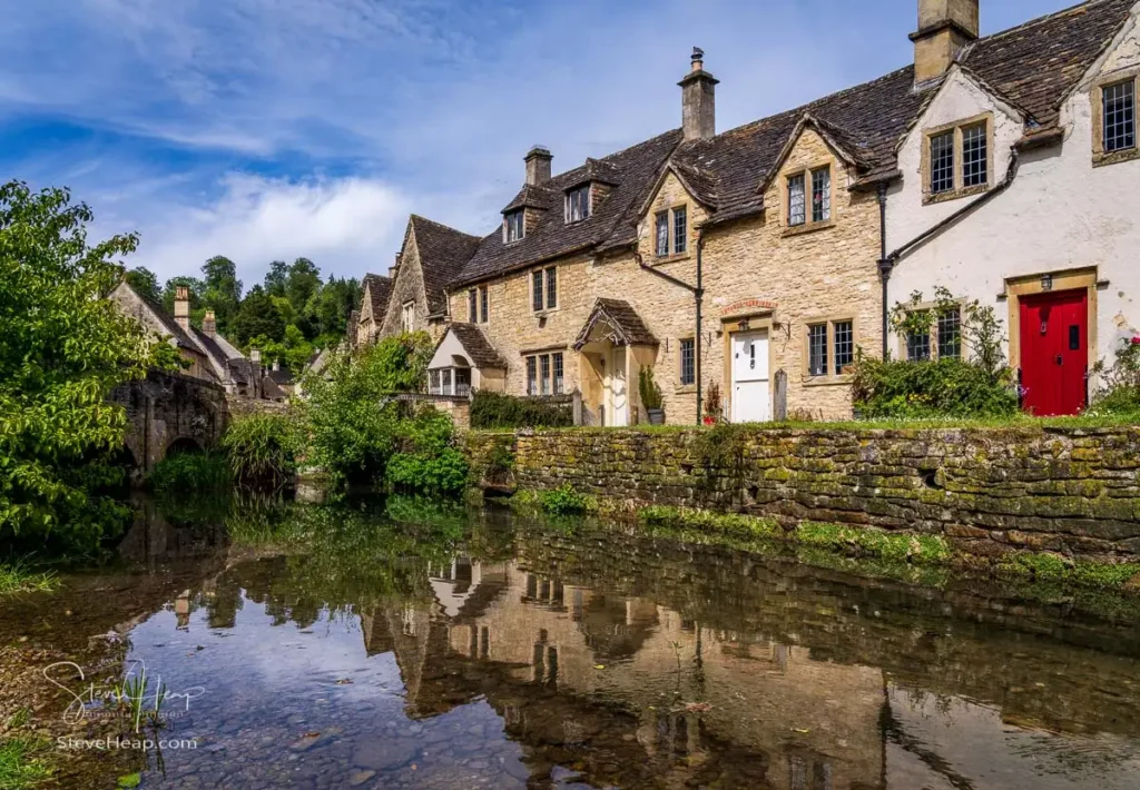 A row of historic honey-colored Cotswold stone cottages reflecting clearly in the calm, still water of a river foreground under a bright sky in Castle Combe, Wiltshire.