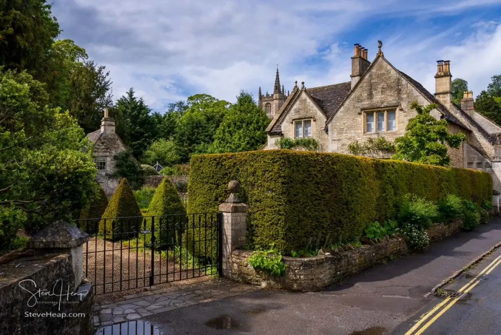 A traditional stone cottage in Castle Combe behind a perfectly manicured green hedge, a stone pillar, and an open black wrought iron gate