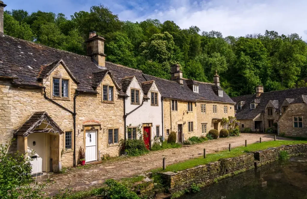 Close-up view of a row of traditional Cotswold stone cottages with tiled roofs and dormer windows, situated below a steep green wooded hillside