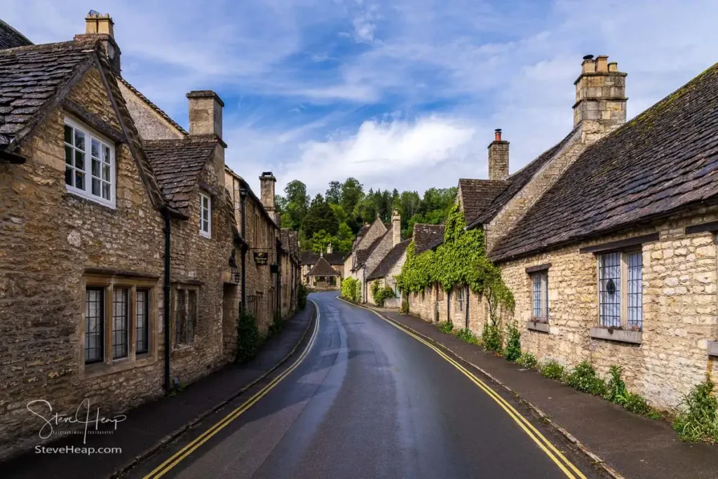 Looking down a narrow, damp asphalt road winding between historic Cotswold stone cottages under a soft, bright sky
