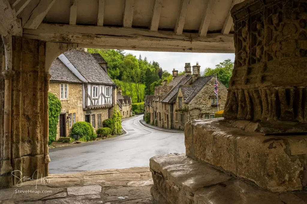 The historic 14th-century stone and timber-roofed Market Cross structure sitting on a raised stone platform in the center of the village square in Castle Combe, Wiltshire.