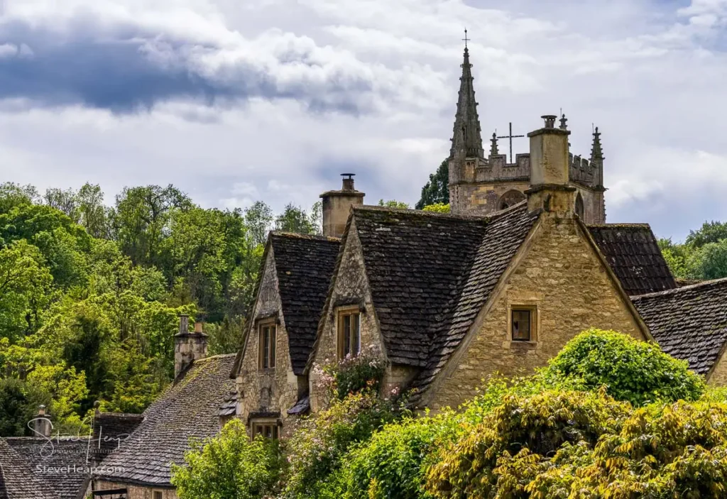 Looking down a village lane heavily framed by deep green trees toward the stone tower of St Andrew's Church and old cottages in Castle Combe