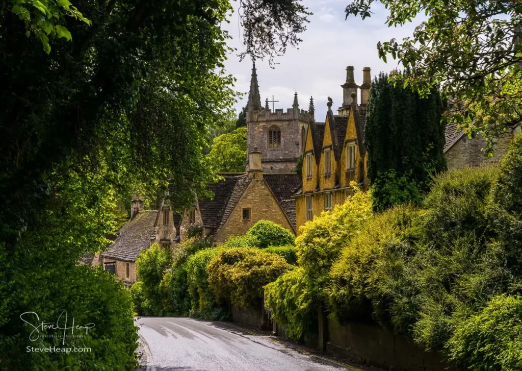 Looking down a village lane heavily framed by deep green trees toward the stone tower of St Andrew's Church and old cottages in Castle Combe.